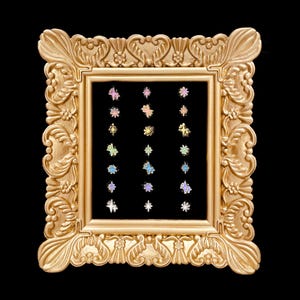 May include: A collection of twenty-one small star-shaped enamel pins arranged in three vertical columns inside an ornate, rectangular gold-colored frame. The pins feature various pastel and metallic colors, including pink, blue, yellow, and silver.