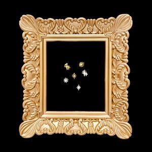 May include: A set of five small star-shaped metal pins in gold and silver tones, arranged in a cluster within a rectangular, ornate, gold-colored picture frame with intricate floral and scrollwork detailing along the border.