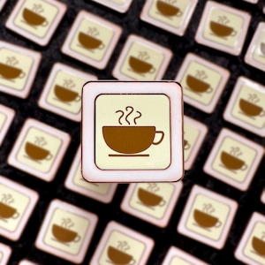 May include: A close-up of a coffee cup pin with a white background and a brown outline. The pin is surrounded by other coffee cup pins, all of which are blurred in the background.