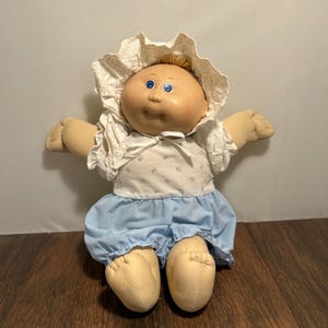 Può includere: Una bambola Cabbage Patch Kid vintage con un viso in vinile morbido e occhi blu. La bambola indossa un cappello bianco con finiture in pizzo, un top bianco con stampa floreale blu e una gonna blu a balze. Le braccia e le gambe della bambola sono in tessuto morbido.
