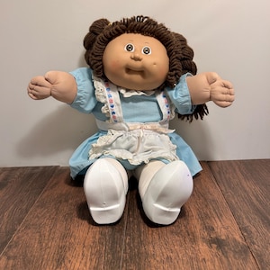 May include: A doll with dark brown yarn hair styled in pigtails, featuring a round face with dimples. The doll wears a light blue dress with a white pinafore apron adorned with lace trim and small floral accents, paired with white shoes and tights.