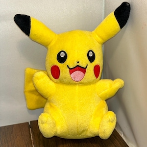 May include: A plush Pikachu toy in bright yellow with black-tipped ears, red cheeks, and a smiling pink mouth. The toy has outstretched arms and is sitting down. The Pikachu toy is a popular character from the Pokemon franchise.