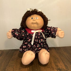 Könnte beinhalten: Eine Vintage-Puppe mit rundem Gesicht, braunen Haaren und einer weißen Schleife. Die Puppe trägt ein dunkles Kleid mit rosa Blumenmustern, einen roten Kragen und ein weißes Band. Arme und Beine der Puppe sind aus weichem, hellem Material.