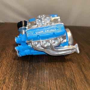 May include: A detailed model of an engine, featuring a vibrant blue body and silver exhaust pipes. The text "Gison Engines" is visible on the engine block. The model is set against a dark wooden surface.