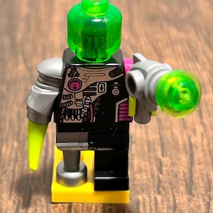 May include: A Lego minifigure with a green transparent head and a black torso with pink and silver details. The figure has a silver arm and a green ray gun. It stands on a yellow base.
