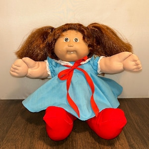May include: A vintage doll with auburn hair styled in pigtails. The doll has a light skin tone, round cheeks, and a small mouth. It wears a light blue dress with a red ribbon and red pants. The doll's arms are outstretched.