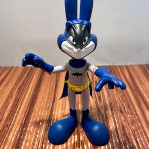 May include: A blue and white Bugs Bunny action figure dressed as Batman. The figure has a black mask, a yellow utility belt, and a gray cape.