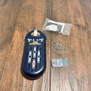 May include: A Star Wars CommTech device in navy blue with white and orange accents. Includes a silver chain, a small clear plastic piece, and a sealed packet. The device has the Star Wars logo.