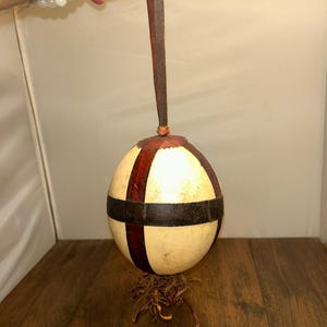 Op de afbeelding: Een bolvormige kalebas-rammelaar met een glad, crèmekleurig oppervlak dat is verdeeld door donkerbruine leren stroken. Een lange, dunne leren riem is aan de bovenkant bevestigd om op te hangen, met onderaan een bundel dunne, gerafelde leren stroken.