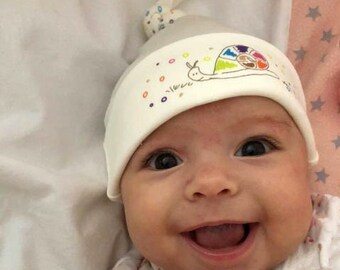 May include: A baby wearing a white hat with a colorful snail design and a red bracelet. The baby is smiling and has their hands up.