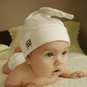 Puede incluir: Un bebé con un gorro blanco de punto con un personaje de dibujos animados marrón en él. El gorro tiene una solapa larga que está doblada sobre la parte superior de la cabeza. El bebé está acostado boca abajo y mirando hacia la derecha.