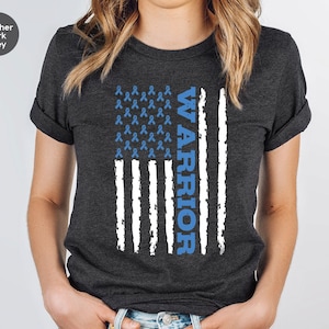 Peut inclure: T-shirt gris foncé chiné avec un motif de drapeau américain blanc et bleu. Le drapeau est composé de symboles de ruban bleu et le mot "WARRIOR" est écrit verticalement sur le côté droit du drapeau.