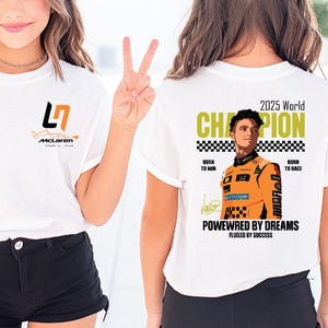 May include: White t-shirt with a McLaren logo on the front and a graphic of a race car driver on the back. The back of the shirt has the text "2025 World Champion" and "Powered by Dreams". The shirt is paired with black shorts.