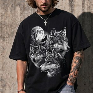 May include: A black short-sleeve t-shirt featuring a graphic print of three wolves and a full moon. The design includes a detailed illustration of wolf heads and a silhouette of a howling wolf against a large, textured lunar background.