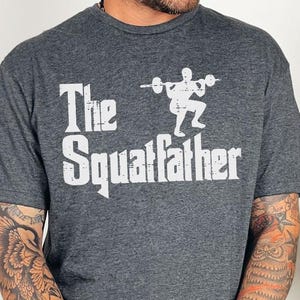 Peut inclure: Un t-shirt gris fonc&eacute; avec un imprim&eacute; graphique blanc d'une personne soulevant des poids avec le texte "The Squatfather".