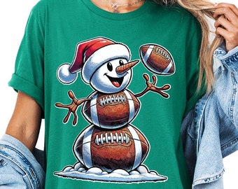 May include: Green t-shirt featuring a snowman made of footballs, wearing a Santa hat and holding a football. The snowman has a carrot nose and twig arms. The design is a festive blend of football and holiday themes.