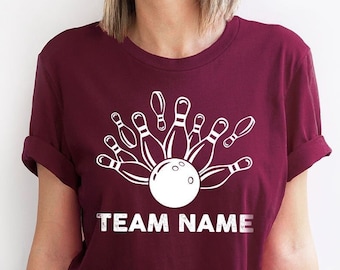 Pode incluir: Camiseta bordô com um design de boliche branco. O gráfico apresenta uma bola de boliche e pinos, com o texto "TEAM NAME" abaixo. A camiseta é de cor sólida e tem mangas enroladas.
