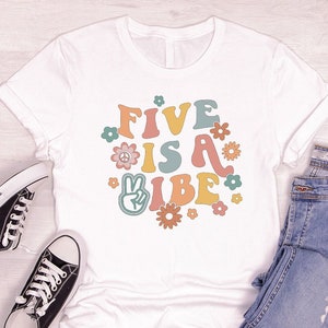 May include: White t-shirt with a graphic design that says "Five is a Vibe" in colorful retro lettering with peace signs and flowers.