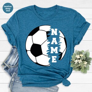 May include: A heather deep teal t-shirt with a white soccer ball graphic. The ball is partially covered by a black and white graphic that says "NAME".