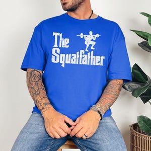 Peut inclure: Un t-shirt bleu royal avec un imprim&eacute; graphique blanc d'une personne accroupie avec une barre et le texte "The Squatfather".