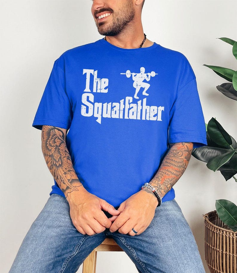 Peut inclure: Un t-shirt bleu royal avec un imprim&eacute; graphique blanc d'une personne accroupie avec une barre et le texte "The Squatfather".