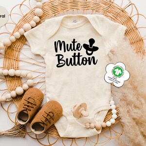 May include: A cream-coloured baby onesie with the words "Mute Button" and a pacifier graphic in black. The onesie is displayed with brown baby shoes, a pacifier, and decorative beads. The background includes a woven wicker placemat and dried pampas grass.