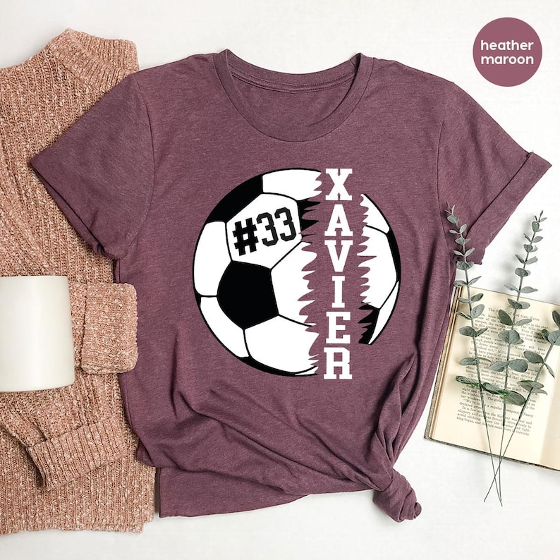 May include: Heather maroon t-shirt featuring a white football graphic with the name "XAVIER" printed vertically down the side of the ball. The football has the number "#33" printed on it.