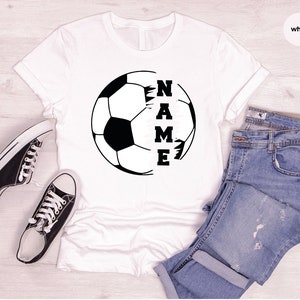 May include: White t-shirt with a black and white soccer ball graphic and the word "NAME" written vertically inside the ball.