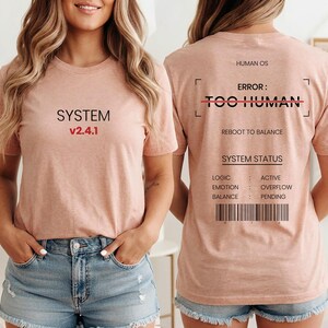 Peut inclure: Un t-shirt couleur pêche avec du texte noir et rouge. L'avant affiche SYSTEM v2.4.1. Le dos présente HUMAN OS, ERROR: TOO HUMAN avec une barre rouge, REBOOT TO BALANCE, SYSTEM STATUS, et une liste pour logic, emotion et balance.