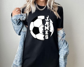 Può includere: Una t-shirt nera a maniche corte con una grande grafica di un pallone da calcio bianco e nero. La parola JAMES è stampata in lettere maiuscole bianche verticalmente al centro del disegno del pallone, che presenta un effetto bordo strappato.
