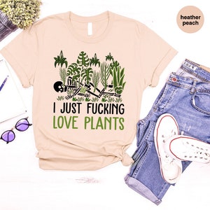 May include: A beige t-shirt with a green graphic of a skeleton surrounded by plants. The text "I JUST FUCKING LOVE PLANTS" is printed in green below the graphic.