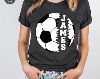 Può includere: Maglietta grigio scuro melange con un grafico di un pallone da calcio bianco. La palla è divisa a metà con il nome "JAMES" scritto verticalmente su un lato.