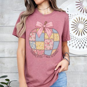 May include: A mauve short-sleeve t-shirt featuring a large pumpkin graphic made of patchwork squares in floral and gingham patterns. A pink bow sits atop the pumpkin, and the text reads TEACHER STYLE BACK TO SCHOOL in a curved arc below.