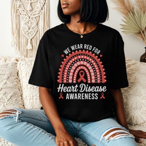 May include: Black t-shirt with a red and white rainbow design and the text "WE WEAR RED FOR Heart Disease AWARENESS". The design includes a red ribbon symbol. The shirt is worn by a person.