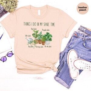 Peut inclure: Un t-shirt beige clair avec le texte "Things I do in my spare time" et un graphique de cinq plantes en pot. Le texte sous le graphique indique "Repot plants", "Propagate plants", "Water plants", "Buy plants", "Rearrange plants", et "Talk with plants".