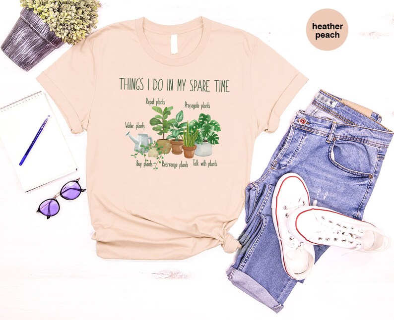 Peut inclure: Un t-shirt beige clair avec le texte "Things I do in my spare time" et un graphique de cinq plantes en pot. Le texte sous le graphique indique "Repot plants", "Propagate plants", "Water plants", "Buy plants", "Rearrange plants", et "Talk with plants".