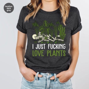 May include: A heather dark grey t-shirt with a skeleton lying in a bed of green plants. The text "I JUST FUCKING LOVE PLANTS" is printed in white below the skeleton.