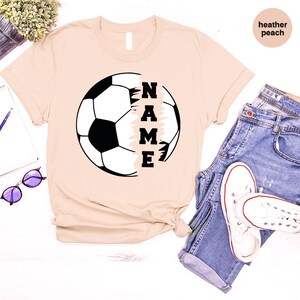May include: A beige t-shirt with a soccer ball graphic and the word "NAME" printed vertically on the right side of the ball. The shirt is made of a soft, comfortable material and is perfect for everyday wear.