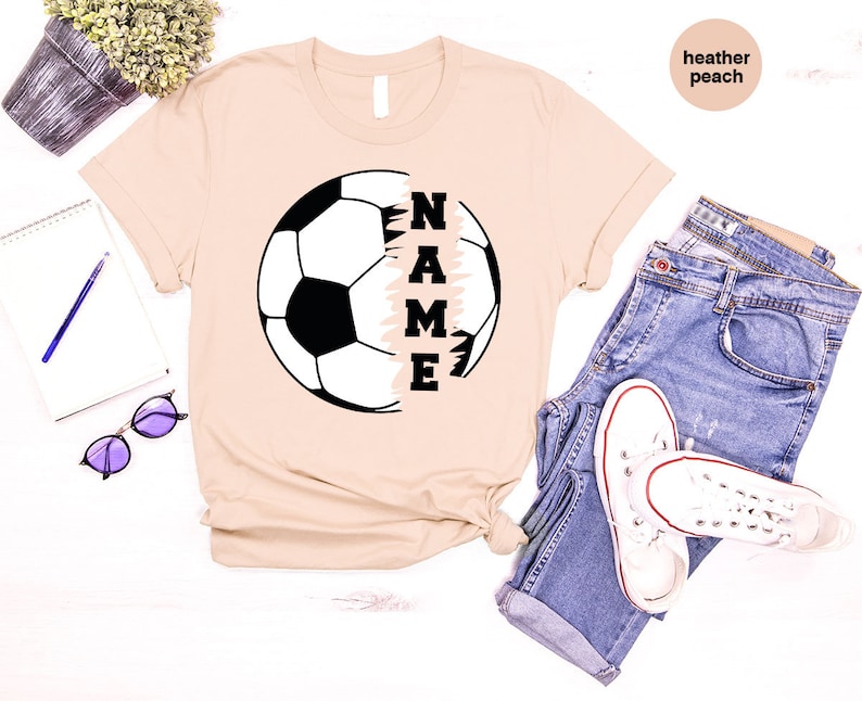 May include: A beige t-shirt with a soccer ball graphic and the word "NAME" printed vertically on the right side of the ball. The shirt is made of a soft, comfortable material and is perfect for everyday wear.