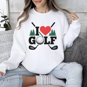 May include: White sweatshirt featuring the text "I LOVE GOLF" in black, a red heart, a golf ball, two golf clubs, and two green pine trees. The sweatshirt has a crew neck and long sleeves.
