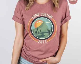 May include: Heather mauve t-shirt with a retro-style graphic of a mountain, trees, and a winding river. The graphic is surrounded by a circle with the text "Camp Custom 2023".