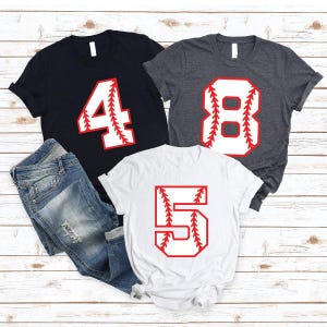 May include: Three short-sleeve t-shirts in black, dark gray, and white, each featuring a large number printed in white with red baseball-style stitching details. The numbers displayed are 4, 8, and 5. A pair of blue denim jeans is included.