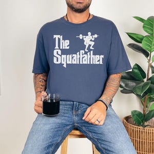 Peut inclure: Un t-shirt bleu marine avec un motif blanc d'une personne soulevant des poids et le texte "The Squatfather".