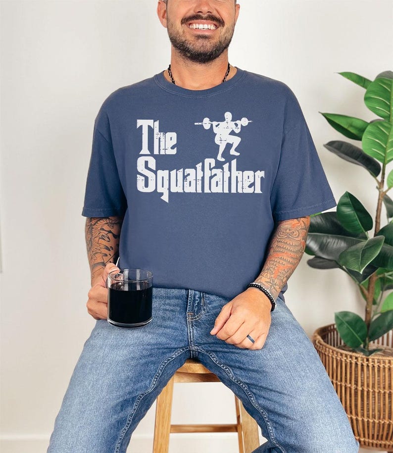 Peut inclure: Un t-shirt bleu marine avec un motif blanc d'une personne soulevant des poids et le texte "The Squatfather".