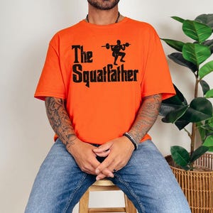 Peut inclure: Un homme portant un t-shirt orange avec le texte "The Squatfather" et une silhouette noire d'une personne soulevant des poids.