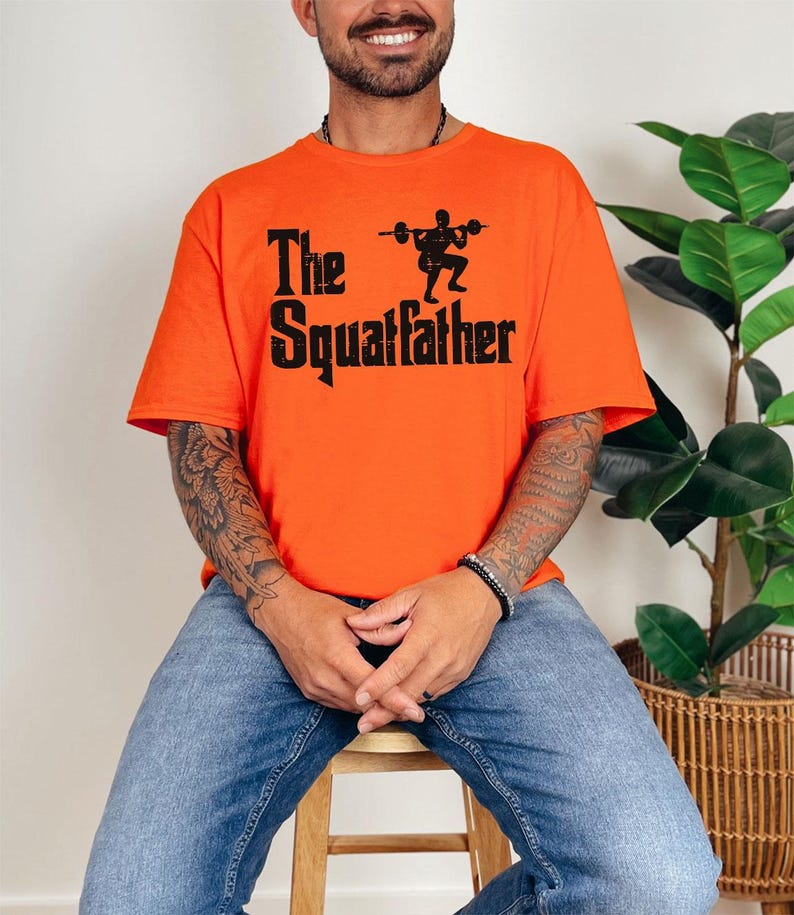 Peut inclure: Un homme portant un t-shirt orange avec le texte "The Squatfather" et une silhouette noire d'une personne soulevant des poids.