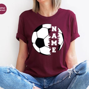 May include: Heather maroon t-shirt with a white soccer ball graphic. The ball is split in half, with the word "NAME" written vertically on the right side.