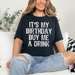 May include: Black t-shirt with the text "IT'S MY BIRTHDAY BUY ME A DRINK" in white. The shirt is worn with light blue jeans. The person is sitting on a white couch with a black and white checkered blanket.