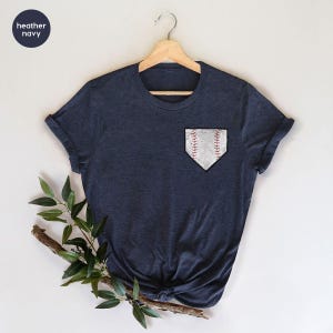 May include: A heather navy short-sleeve t-shirt featuring a graphic on the left chest area. The graphic is a white home plate shape with a textured baseball pattern, including red stitching details. The shirt is displayed on a wooden hanger.
