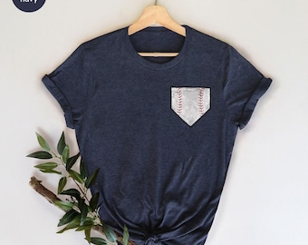 May include: A heather navy short-sleeve t-shirt featuring a graphic on the left chest area. The graphic is a white home plate shape with a textured baseball pattern, including red stitching details. The shirt is displayed on a wooden hanger.